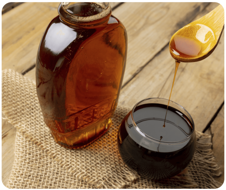 A bottle of maple syrup placed on a wooden table, adjacent to a glass bowl from which a ladle is scooping up the golden syrup, highlighting the rich, amber color of the maple syrup against the rustic backdrop